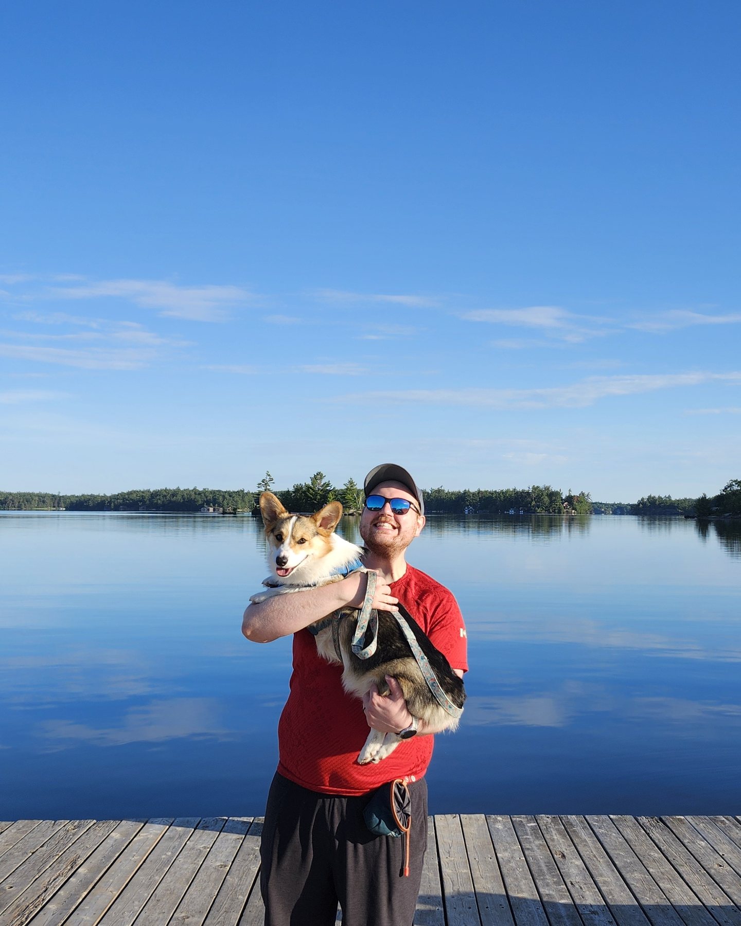 Glen holding Moose, a corgi, on a dock beside a calm Muskoka lake on a clear summer morning